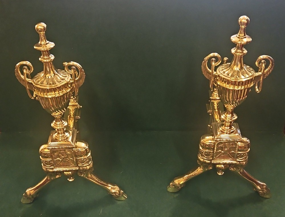 A pair of polished brass Regency fire dogs featuring classical urn finials and splayed support feet.