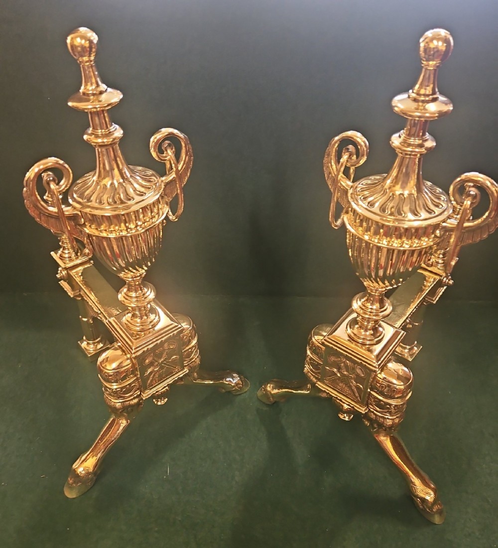 A pair of polished brass Regency fire dogs featuring classical urn finials and splayed support feet. View II