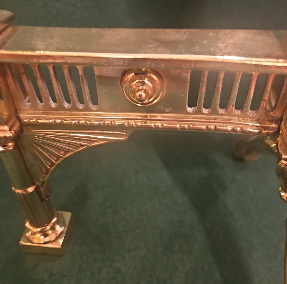 A pair of polished brass Regency fire dogs featuring classical urn finials and splayed support feet. View III