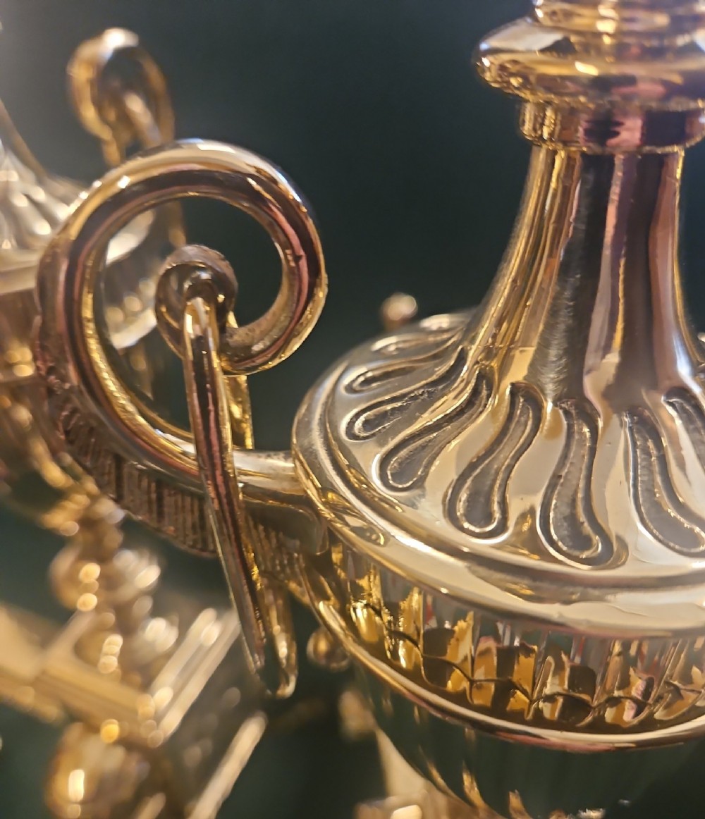 A pair of polished brass Regency fire dogs featuring classical urn finials and splayed support feet. View V