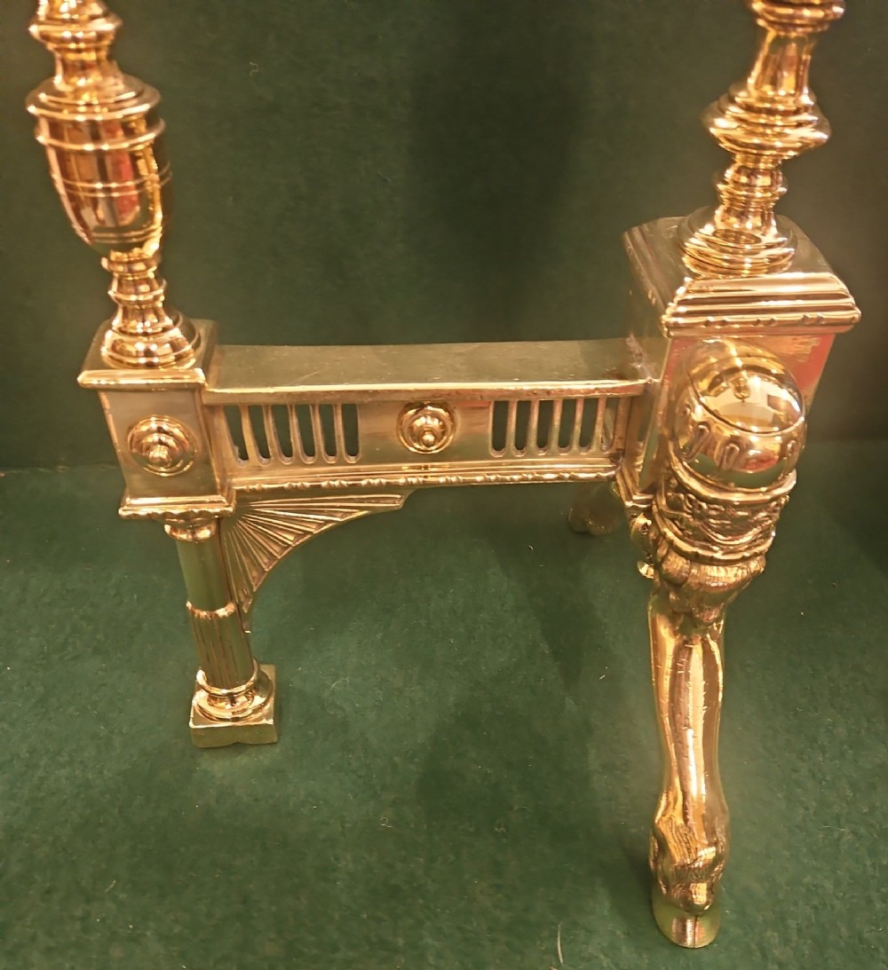 A pair of polished brass Regency fire dogs featuring classical urn finials and splayed support feet. View VII