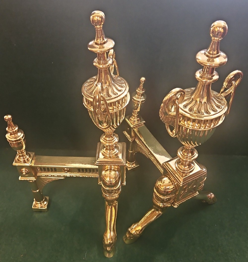 A pair of polished brass Regency fire dogs featuring classical urn finials and splayed support feet. View VIII
