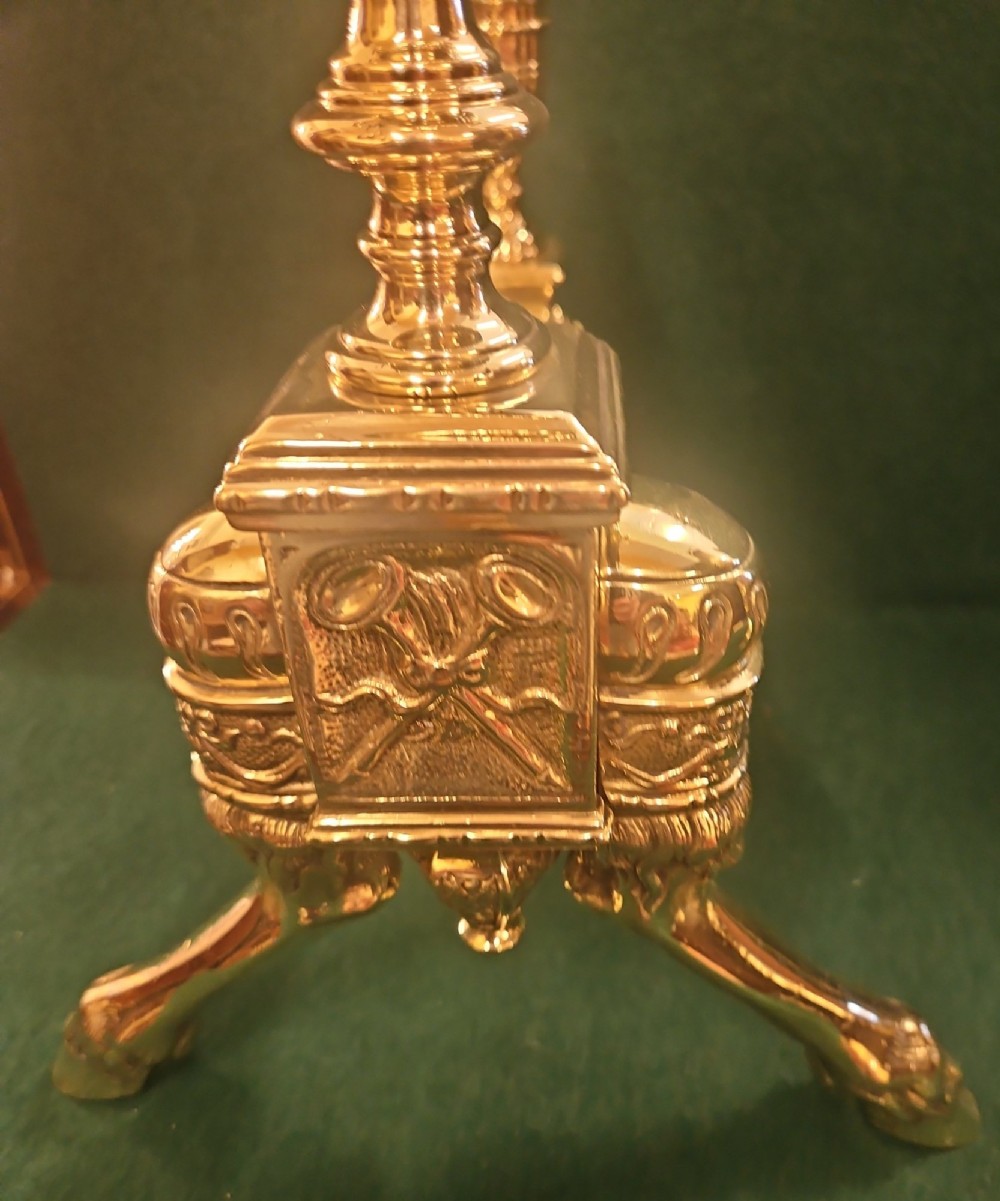 A pair of polished brass Regency fire dogs featuring classical urn finials and splayed support feet. View IX