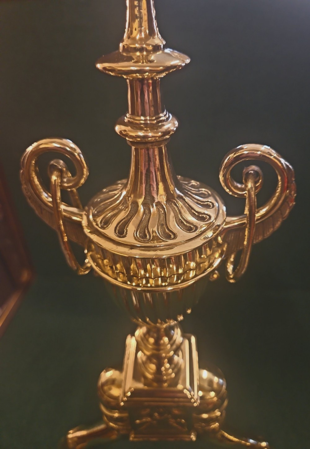 A pair of polished brass Regency fire dogs featuring classical urn finials and splayed support feet. View X