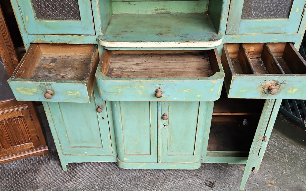 A distressed green painted pine kitchen cabinet featuring upper glazed doors, drawers, and lower panelled wooden doors. View III