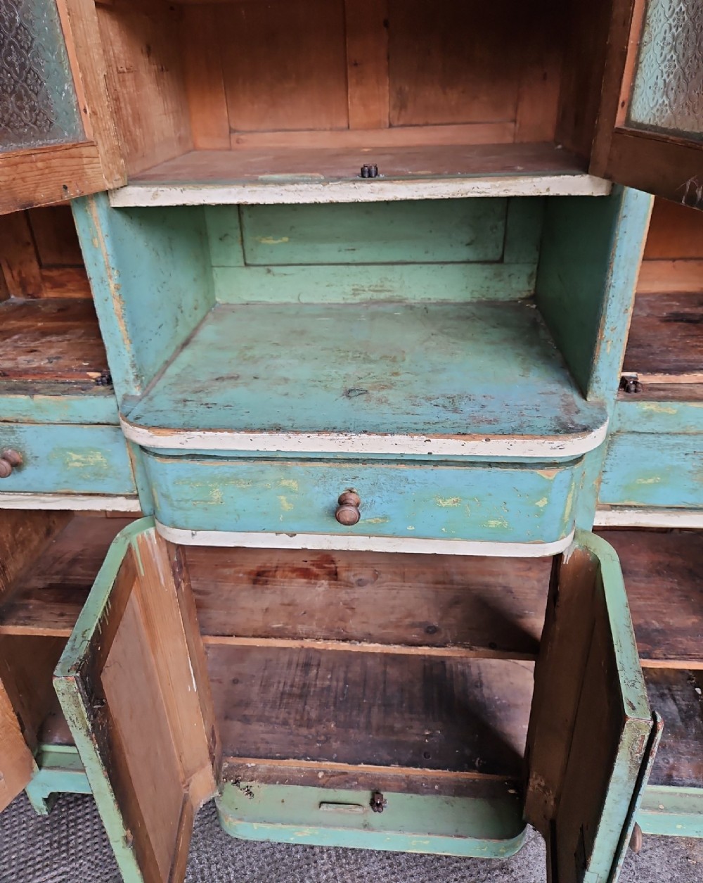 A distressed green painted pine kitchen cabinet featuring upper glazed doors, drawers, and lower panelled wooden doors. View VII