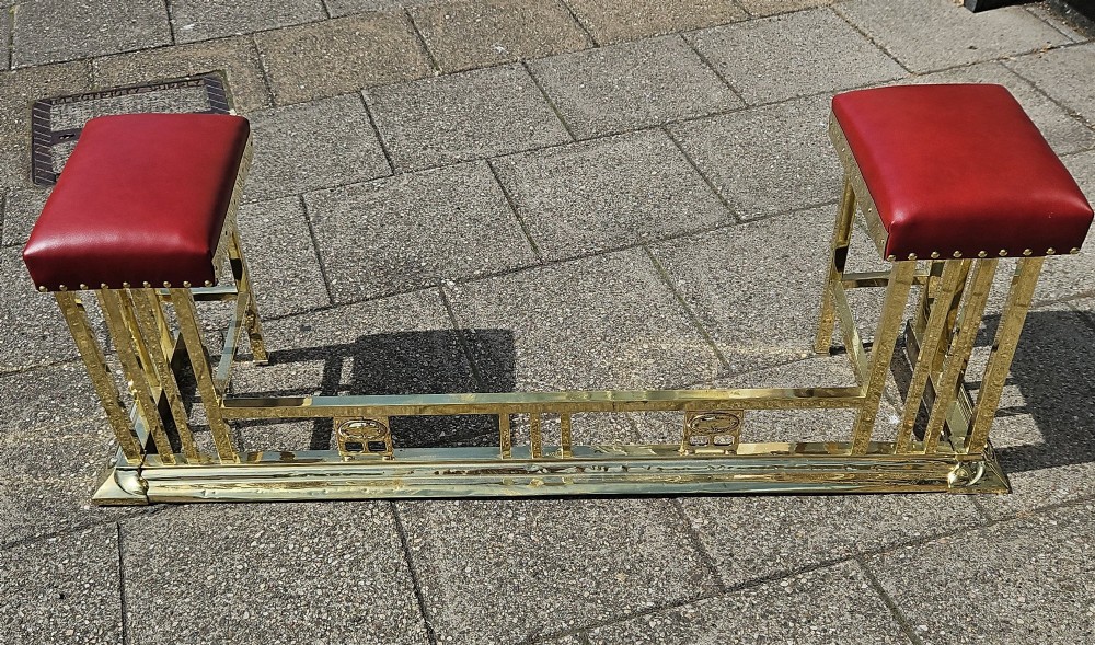 A Victorian brass club fender featuring a polished metal frame with upright supports and two red leather buttoned seats.