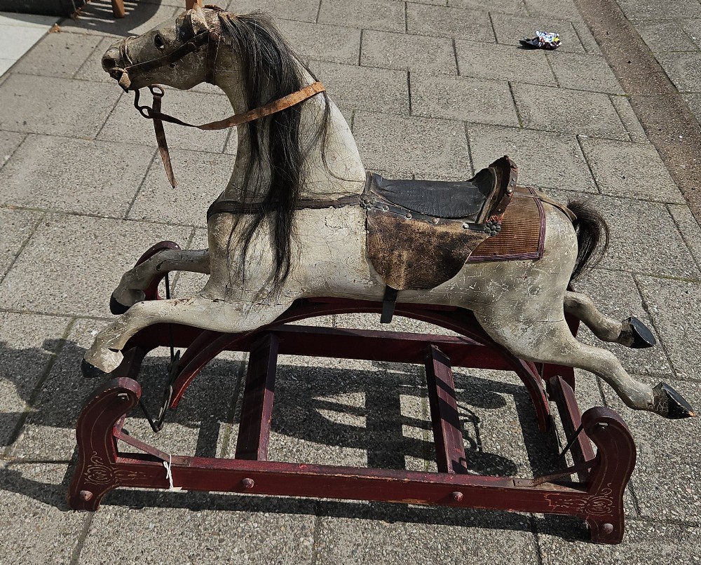 An antique carved wooden rocking horse with a grey painted finish, dark leather saddle, and black horsehair mane, mounted on a dark red glider stand.