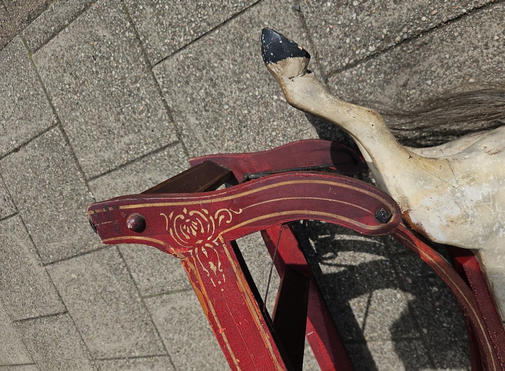 An antique carved wooden rocking horse with a grey painted finish, dark leather saddle, and black horsehair mane, mounted on a dark red glider stand. View III