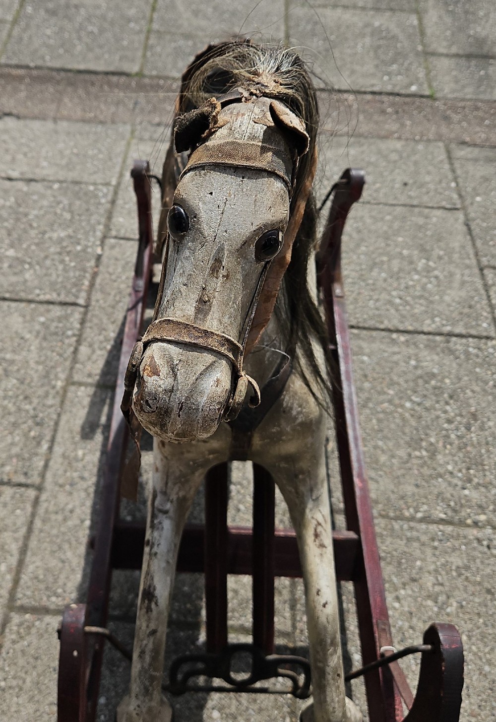 An antique carved wooden rocking horse with a grey painted finish, dark leather saddle, and black horsehair mane, mounted on a dark red glider stand. View VI