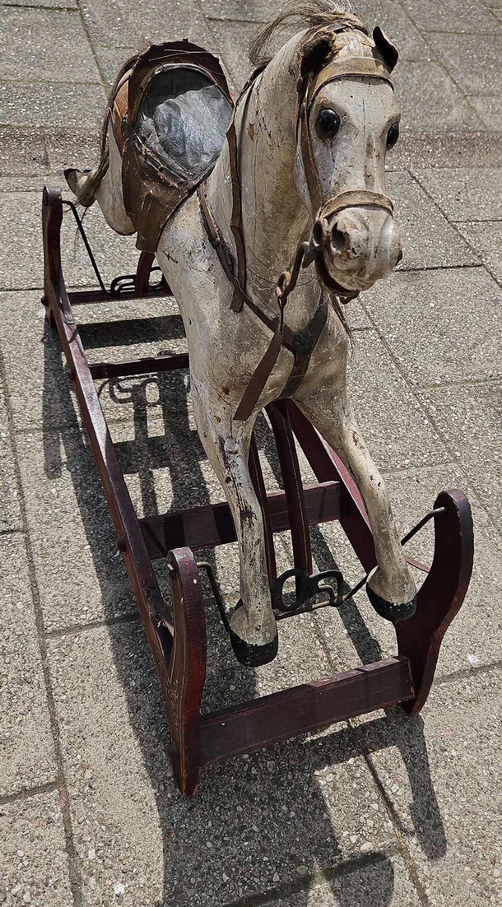 An antique carved wooden rocking horse with a grey painted finish, dark leather saddle, and black horsehair mane, mounted on a dark red glider stand. View VII