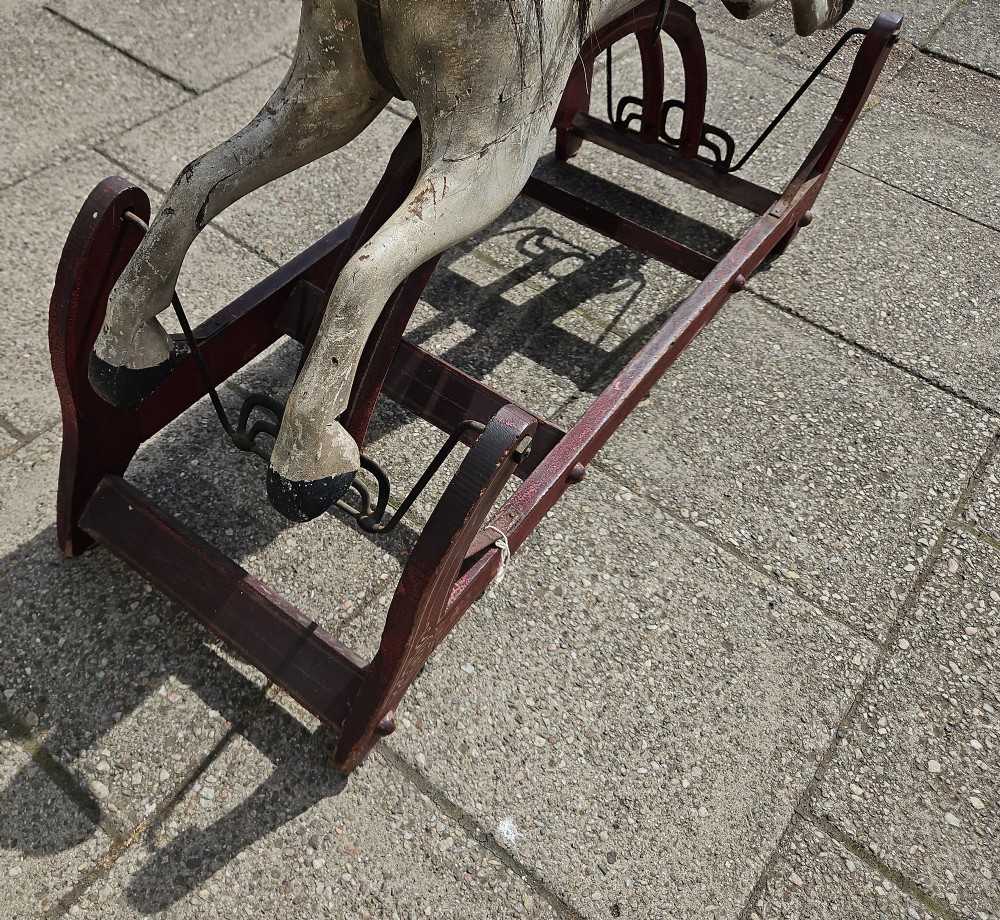 An antique carved wooden rocking horse with a grey painted finish, dark leather saddle, and black horsehair mane, mounted on a dark red glider stand. View X