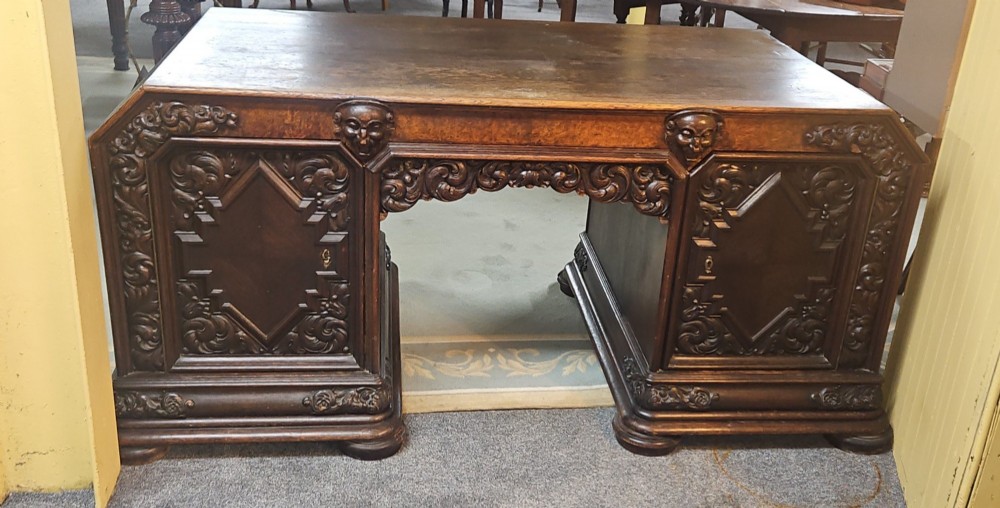 A dark carved oak pedestal desk featuring intricate relief scrollwork, carved mask motifs, geometric diamond side panels, and a flat rectangular top. View VI