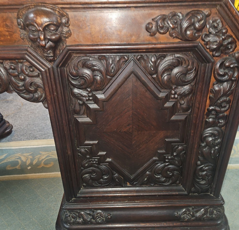 A dark carved oak pedestal desk featuring intricate relief scrollwork, carved mask motifs, geometric diamond side panels, and a flat rectangular top. View IX