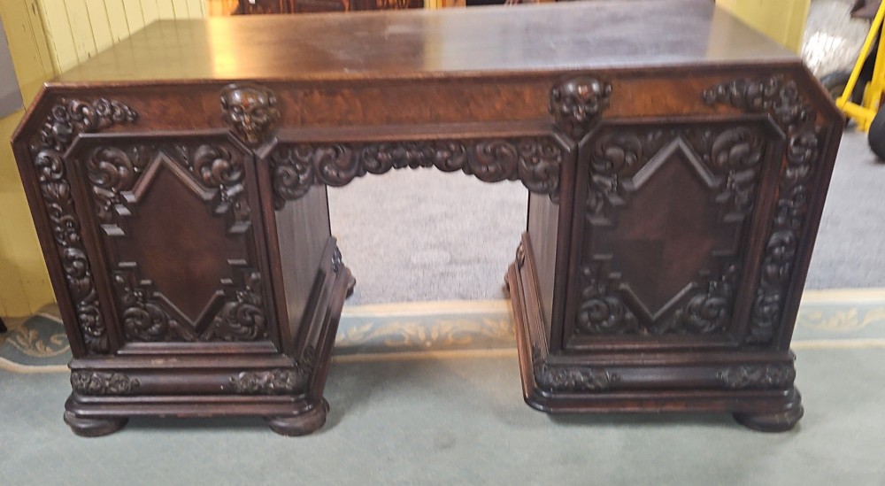 A dark carved oak pedestal desk featuring intricate relief scrollwork, carved mask motifs, geometric diamond side panels, and a flat rectangular top. View X