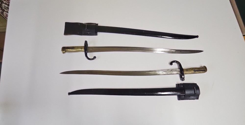 A pair of 19th-century French sword bayonets featuring ribbed brass hilts, recurved quillons, steel blades, black scabbards, and leather frogs on a plain surface. View II
