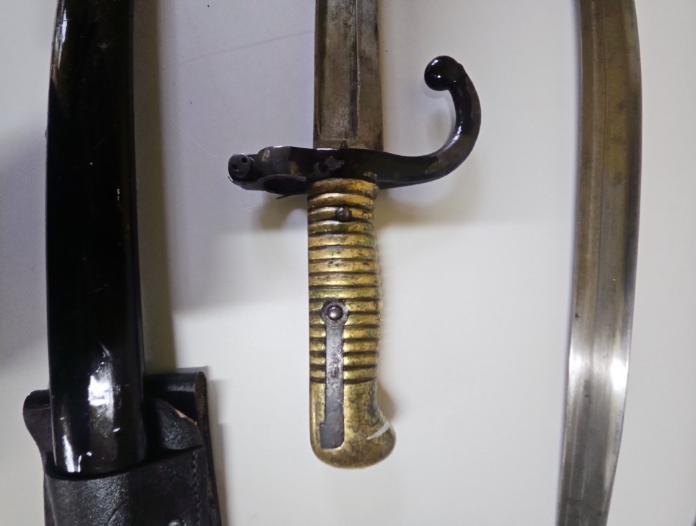A pair of 19th-century French sword bayonets featuring ribbed brass hilts, recurved quillons, steel blades, black scabbards, and leather frogs on a plain surface. View III