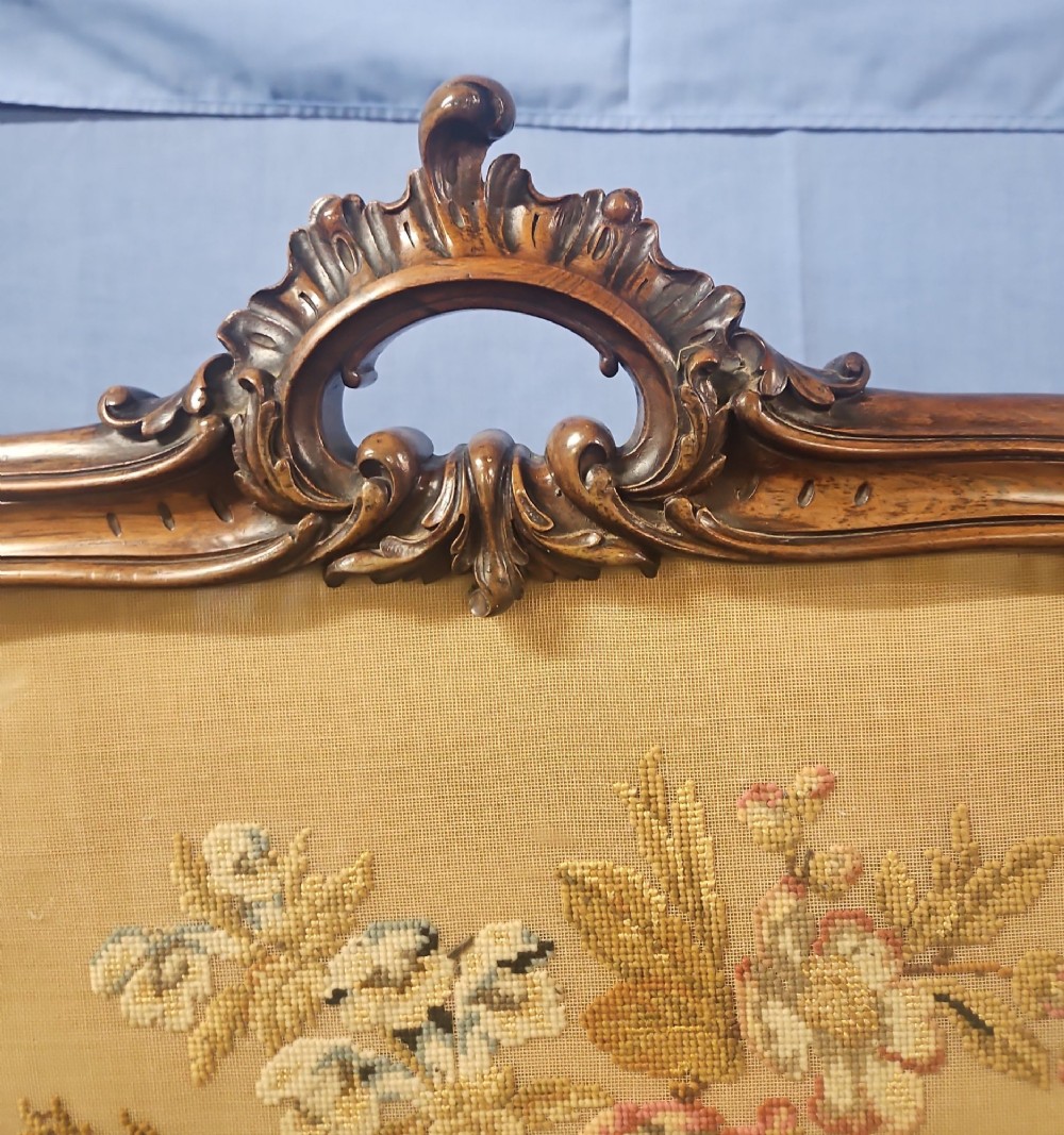 A Victorian carved walnut fire screen featuring an ornate cresting frame supported by cabriole legs, encasing a floral needlepoint textile panel. View IV