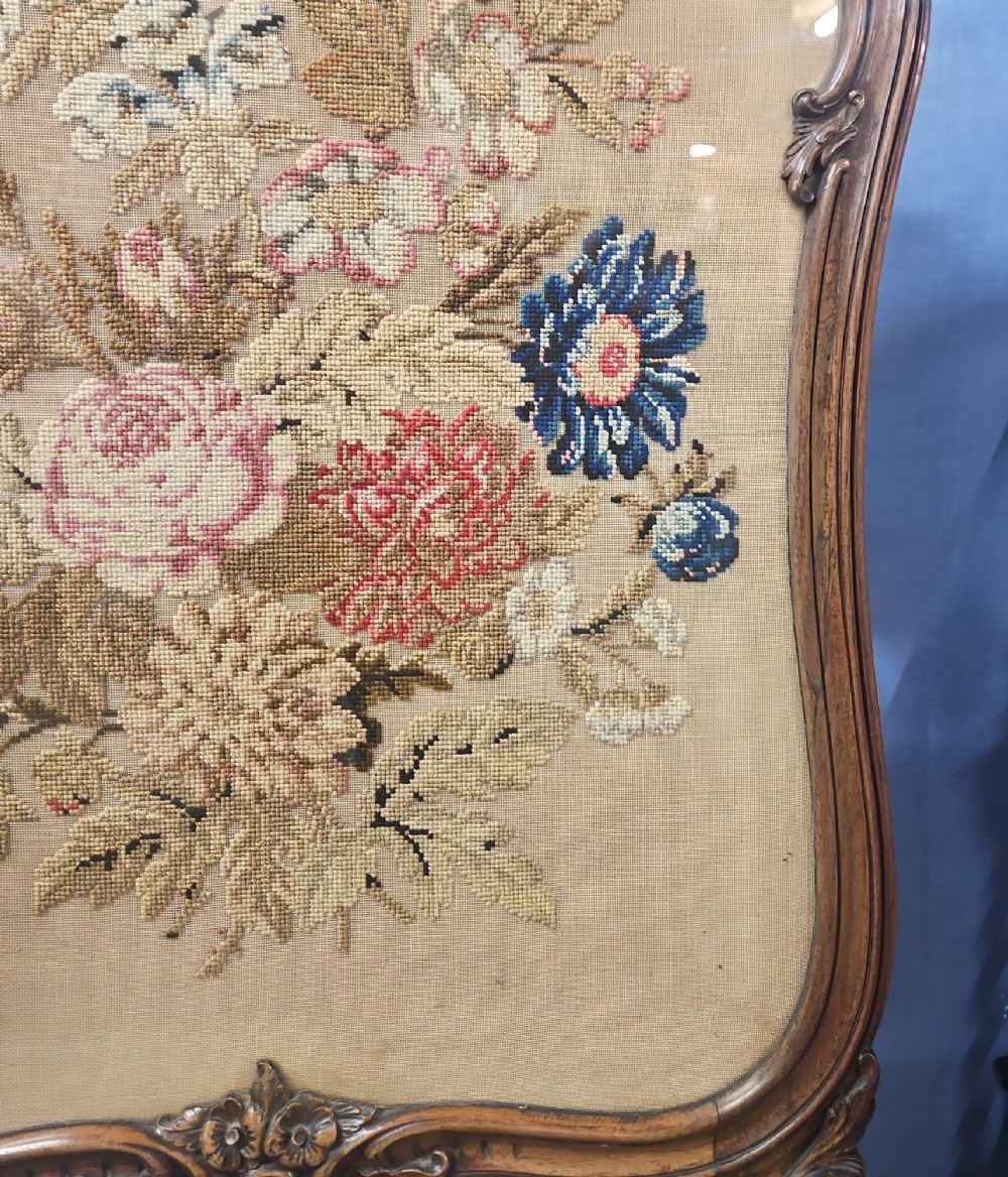 A Victorian carved walnut fire screen featuring an ornate cresting frame supported by cabriole legs, encasing a floral needlepoint textile panel. View VIII
