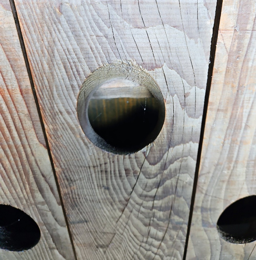 A vertical weathered timber champagne riddling rack featuring multiple angled circular cutouts and stamped with Champagne Mumm Reims France near the base. View II