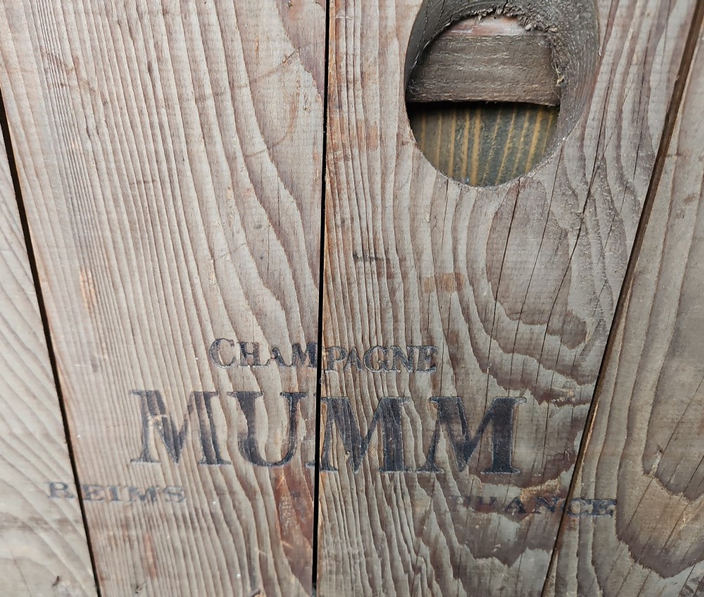 A vertical weathered timber champagne riddling rack featuring multiple angled circular cutouts and stamped with Champagne Mumm Reims France near the base. View IV
