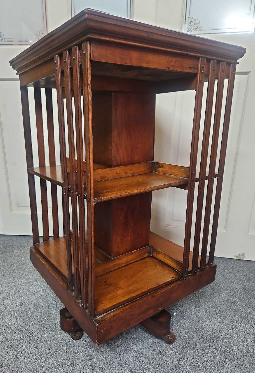 An Edwardian mahogany revolving bookcase featuring vertical slatted sides, two tiers of open shelving, and a square moulded top set on a rotating cross base.