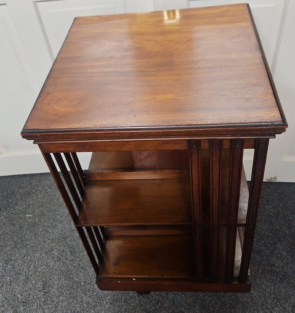 An Edwardian mahogany revolving bookcase featuring vertical slatted sides, two tiers of open shelving, and a square moulded top set on a rotating cross base. View IV