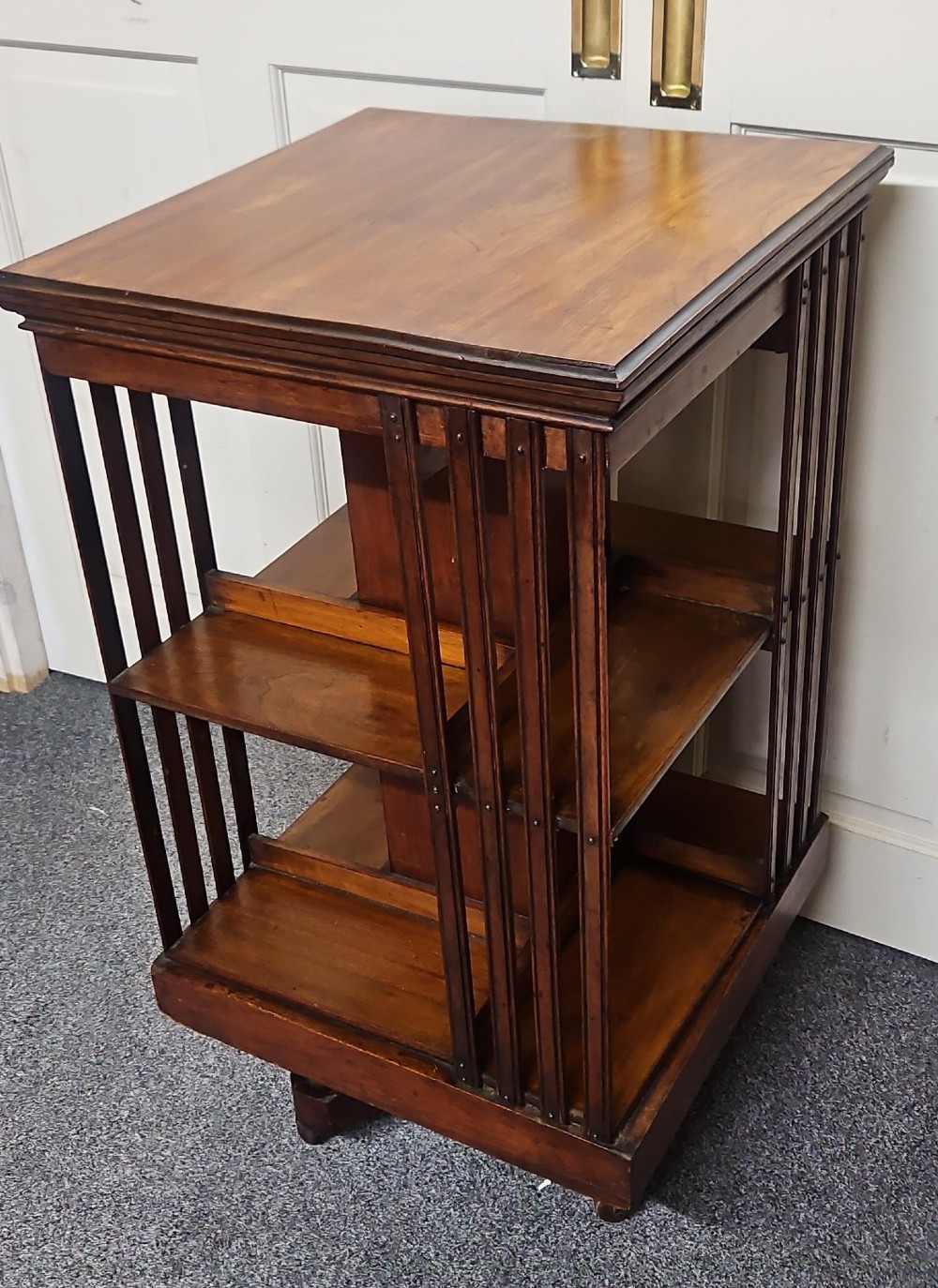 An Edwardian mahogany revolving bookcase featuring vertical slatted sides, two tiers of open shelving, and a square moulded top set on a rotating cross base. View VI