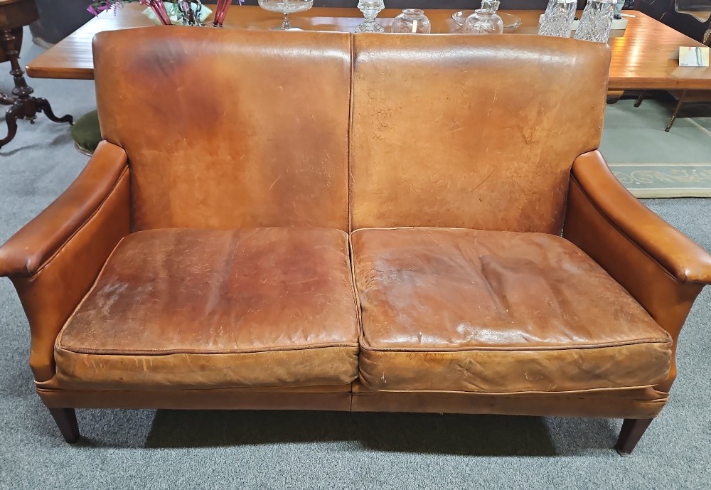 An Edwardian warm brown leather sofa featuring aged surface patina, loose seat cushions, and short tapered wooden legs.