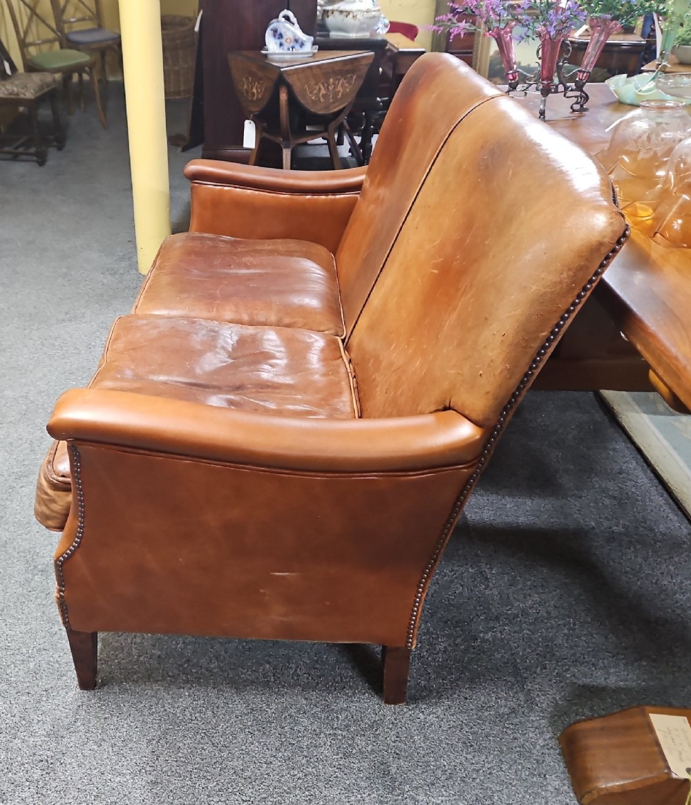 An Edwardian warm brown leather sofa featuring aged surface patina, loose seat cushions, and short tapered wooden legs. View V