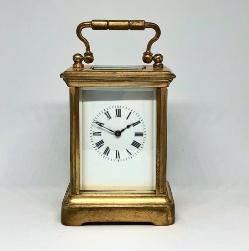 fig 1. A late nineteenth-century miniature French gilt brass carriage clock featuring a rectangular case with glass panels, a curved top handle, and a white enamel dial with Roman numerals.