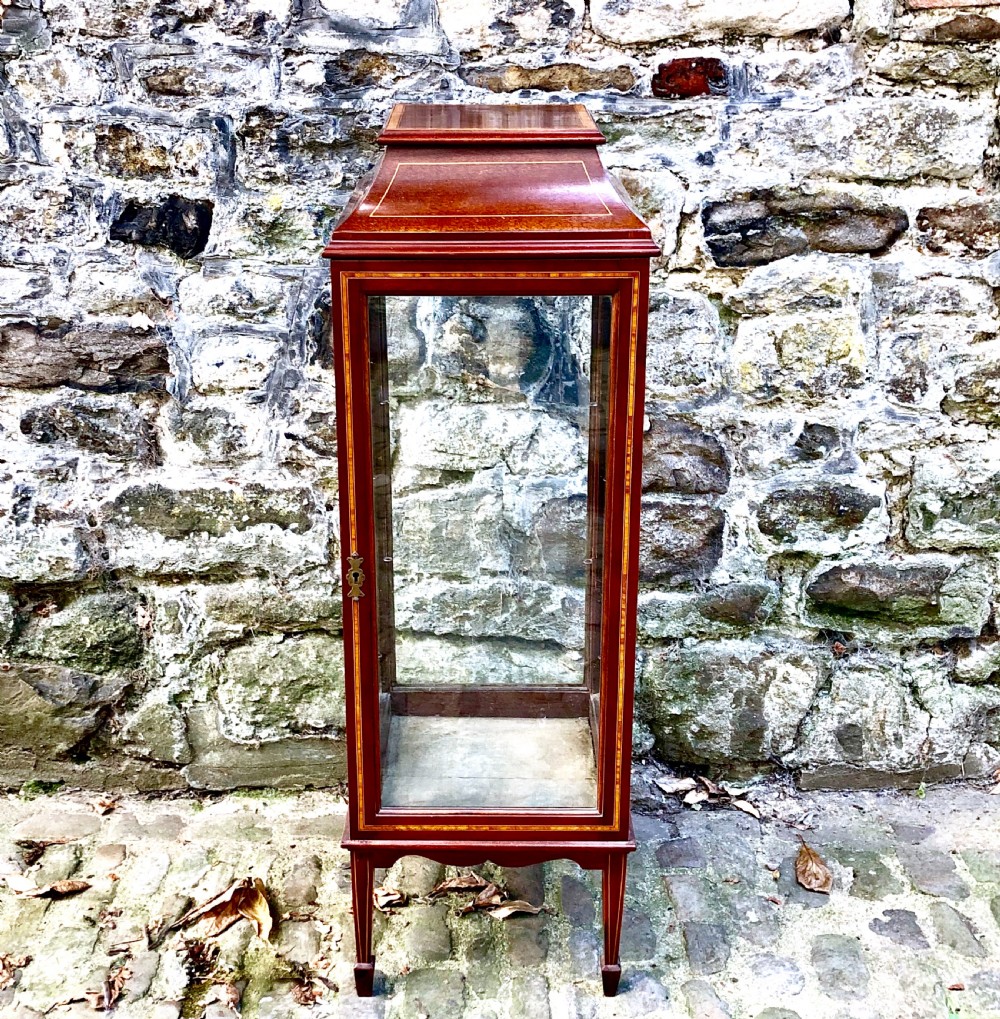 A tall, narrow Edwardian mahogany display cabinet with glazed side panels, fine wood stringing inlay, a single glass front door with a brass keyhole, and square tapered legs.
