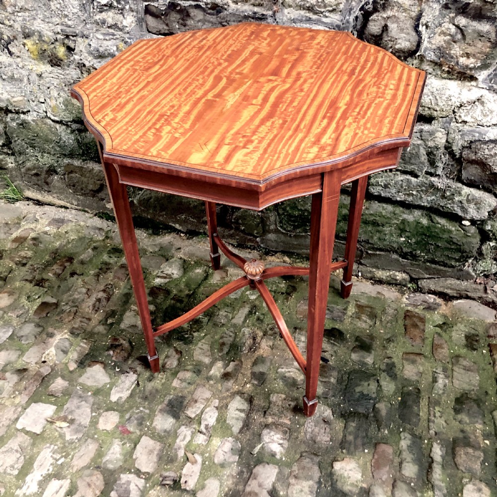An Edwardian satinwood occasional table with a shaped top, crossbanded edging, square tapered legs, and an X-frame stretcher with a central finial. View II