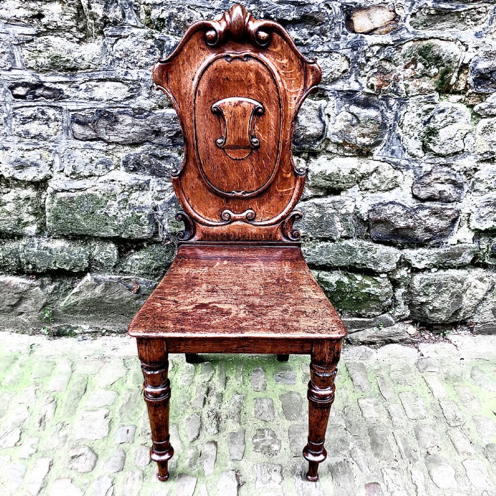 An early Victorian oak hall chair featuring a carved shield-shaped backrest, solid wooden seat, and turned front legs.
