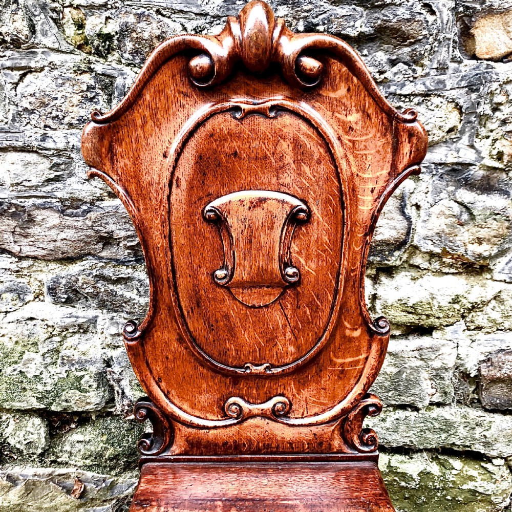 An early Victorian oak hall chair featuring a carved shield-shaped backrest, solid wooden seat, and turned front legs. View V