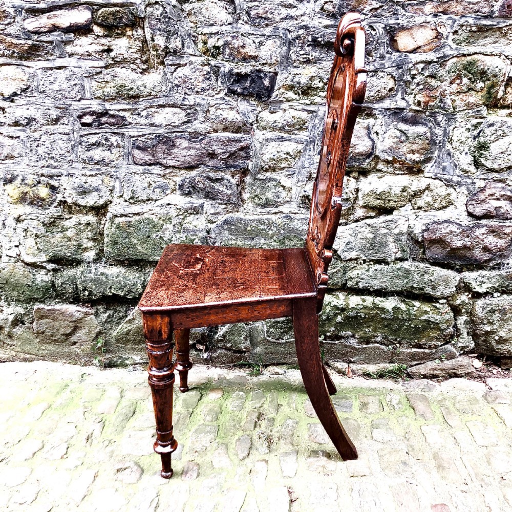 An early Victorian oak hall chair featuring a carved shield-shaped backrest, solid wooden seat, and turned front legs. View VI