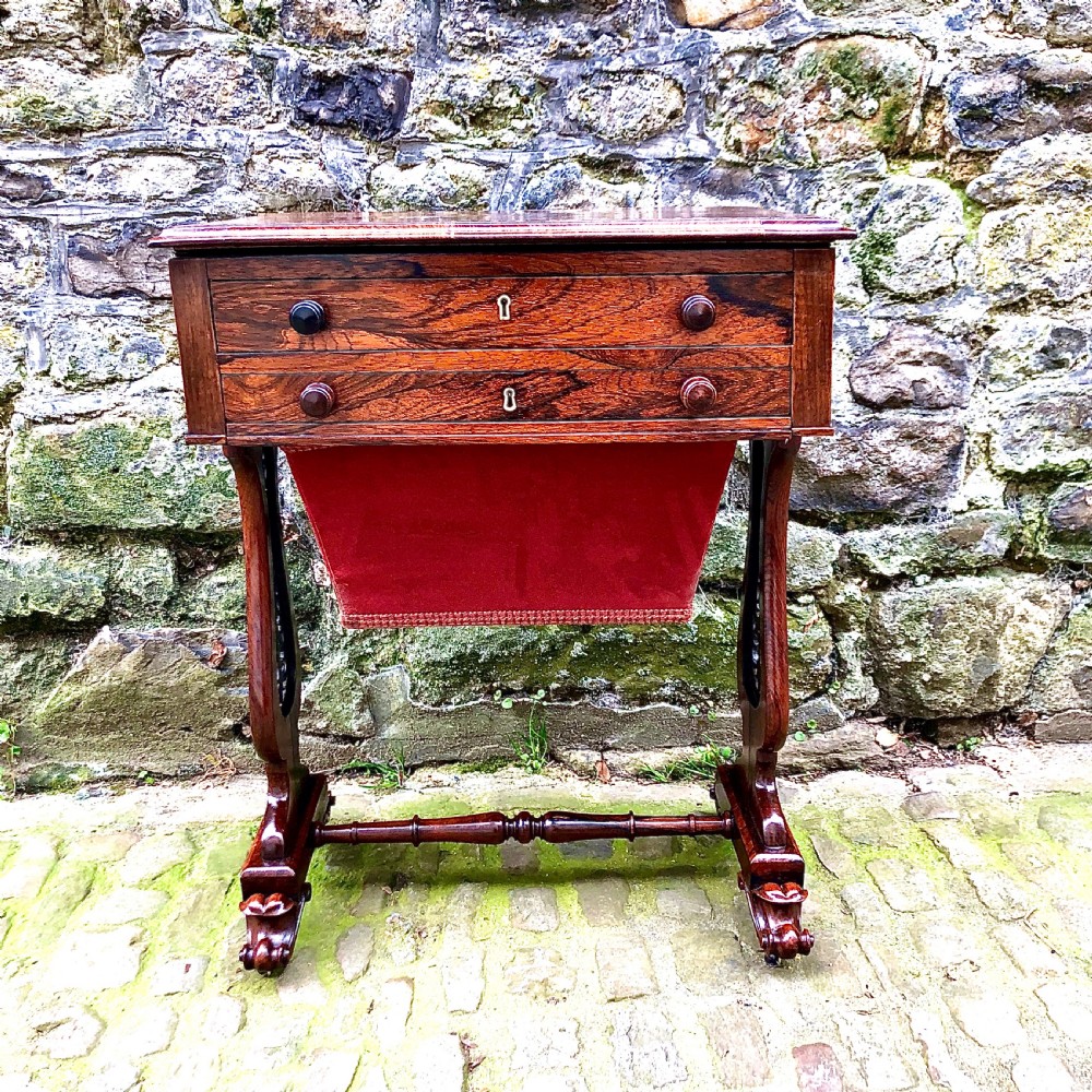 A William IV rosewood work table featuring two drawer fronts, turned knobs, keyhole escutcheons, a red fabric drop pouch, and carved end supports.