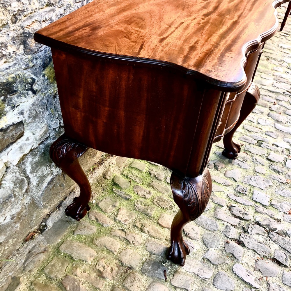 A Portuguese mahogany two-drawer commode featuring serpentine drawer fronts, decorative brass escutcheons and handles, and carved cabriole legs ending in claw feet. View VI