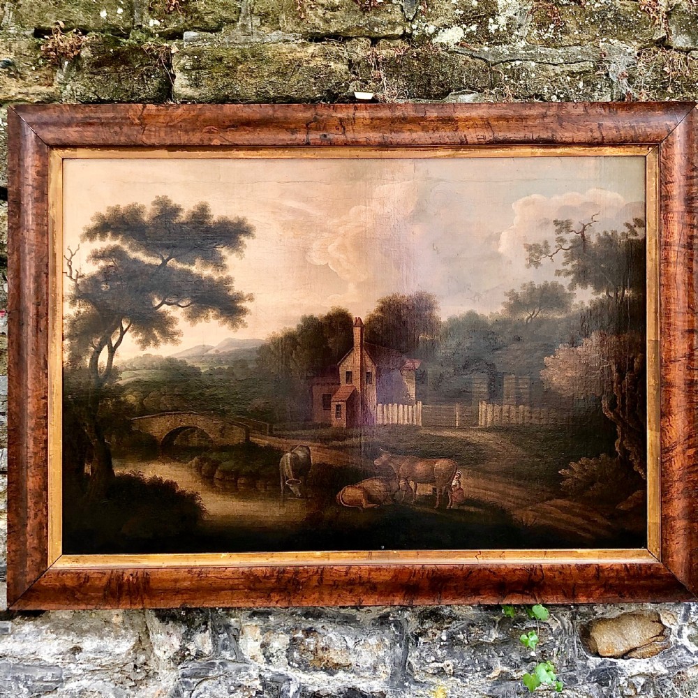 An early nineteenth-century naive oil painting depicting a pastoral scene with cattle, a stone arch bridge, and a rural cottage, housed in a wide timber frame with a gold leaf inner slip.