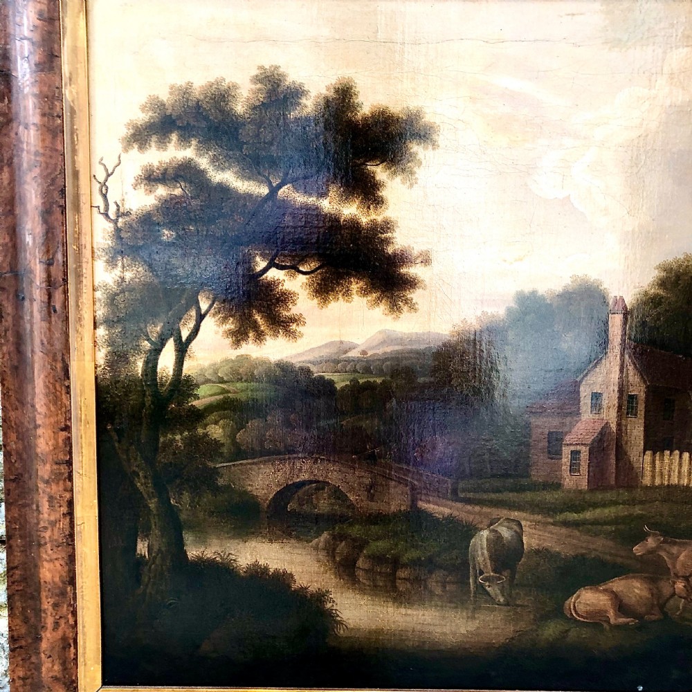 An early nineteenth-century naive oil painting depicting a pastoral scene with cattle, a stone arch bridge, and a rural cottage, housed in a wide timber frame with a gold leaf inner slip. View III