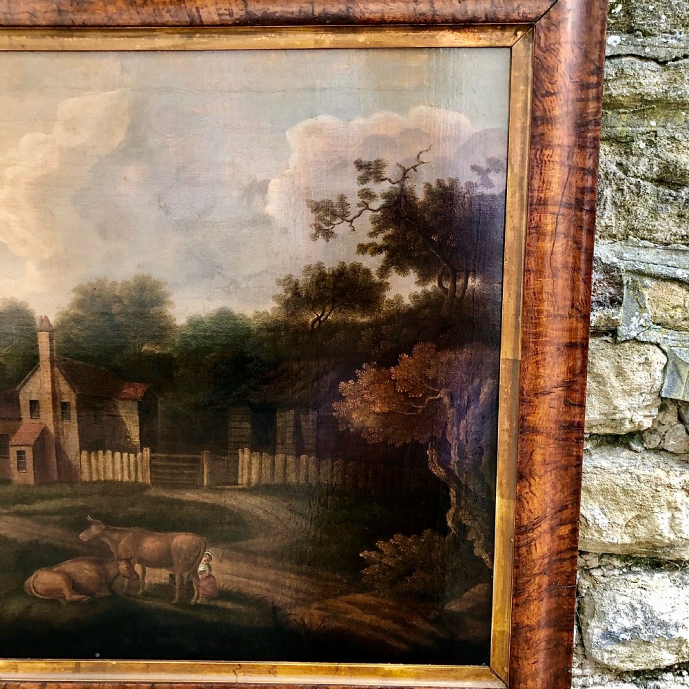 An early nineteenth-century naive oil painting depicting a pastoral scene with cattle, a stone arch bridge, and a rural cottage, housed in a wide timber frame with a gold leaf inner slip. View V
