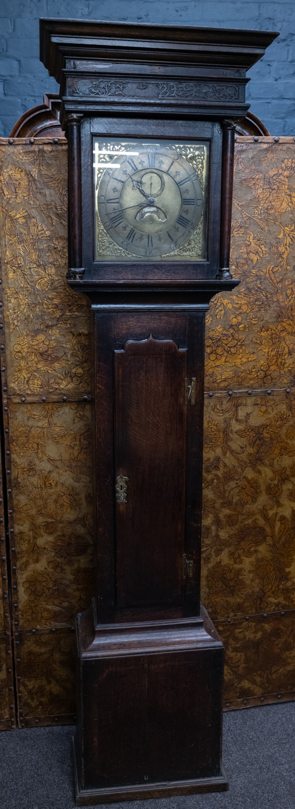 An antique dark oak longcase clock featuring a carved hood, a square brass dial with Roman numerals, and a tall wooden trunk door.