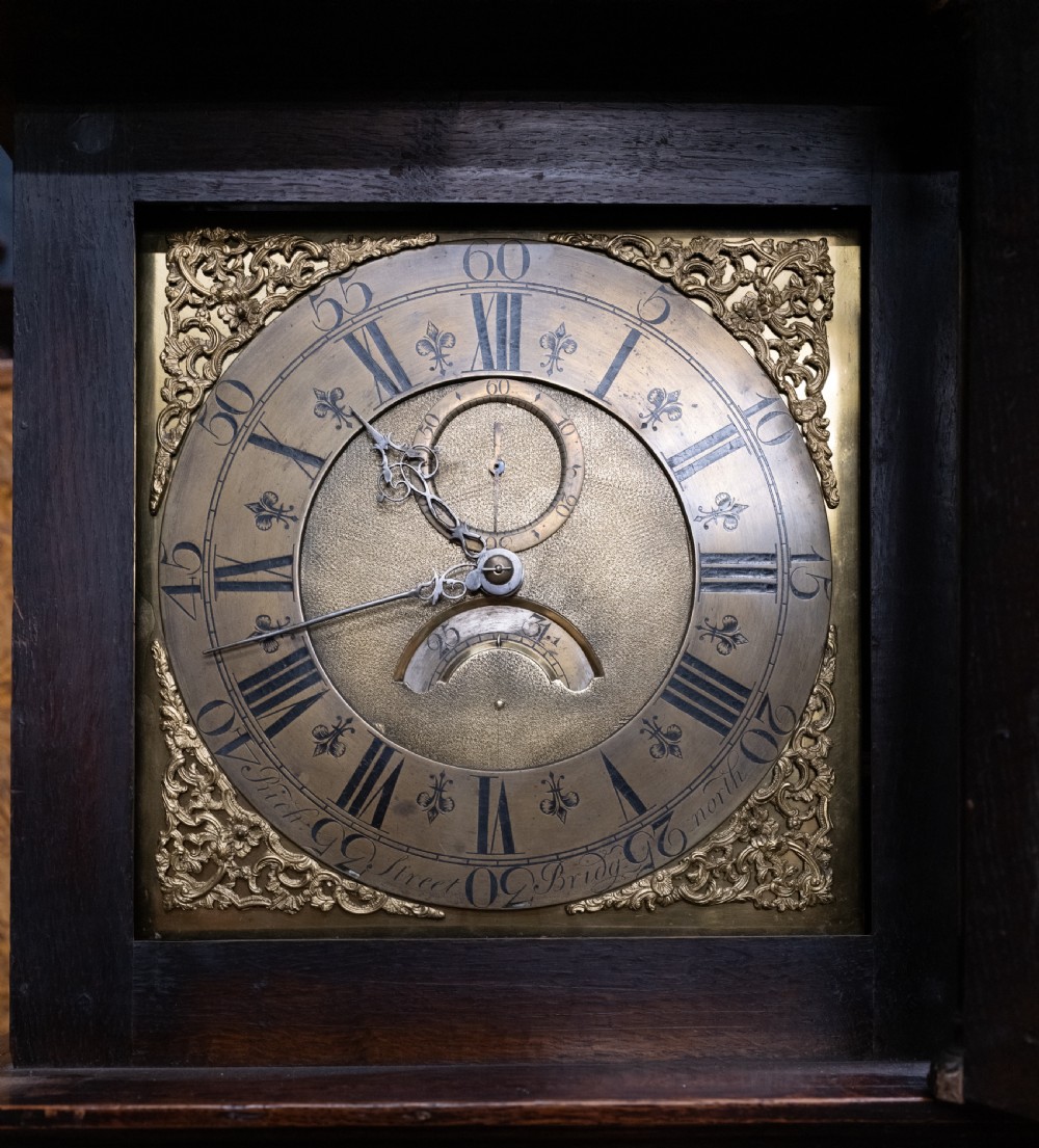 An antique dark oak longcase clock featuring a carved hood, a square brass dial with Roman numerals, and a tall wooden trunk door. View IV