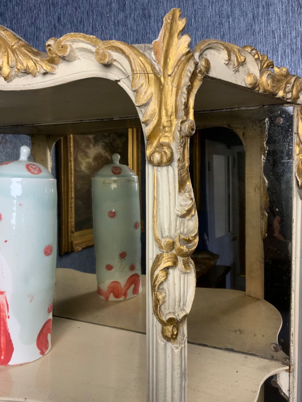 A tall floor-standing cream painted and parcel gilt open display cabinet with four tiers, a mirrored back, and carved Rococo cresting. View III