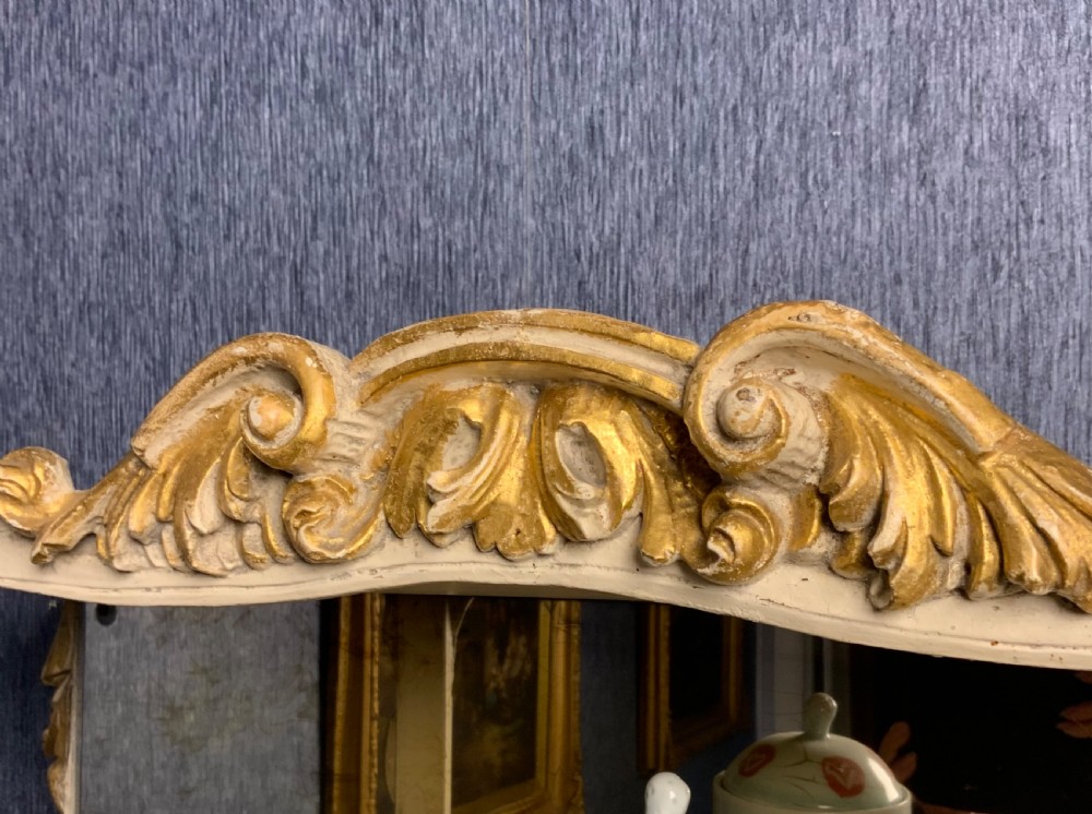 A tall floor-standing cream painted and parcel gilt open display cabinet with four tiers, a mirrored back, and carved Rococo cresting. View V