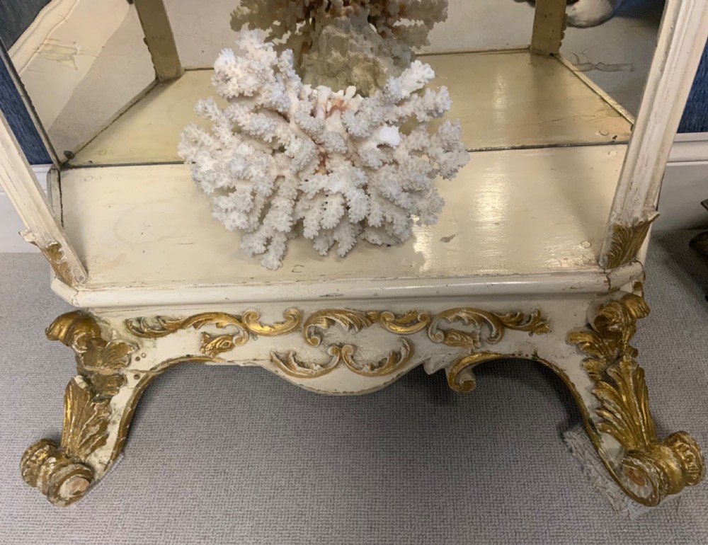 A tall floor-standing cream painted and parcel gilt open display cabinet with four tiers, a mirrored back, and carved Rococo cresting. View VIII