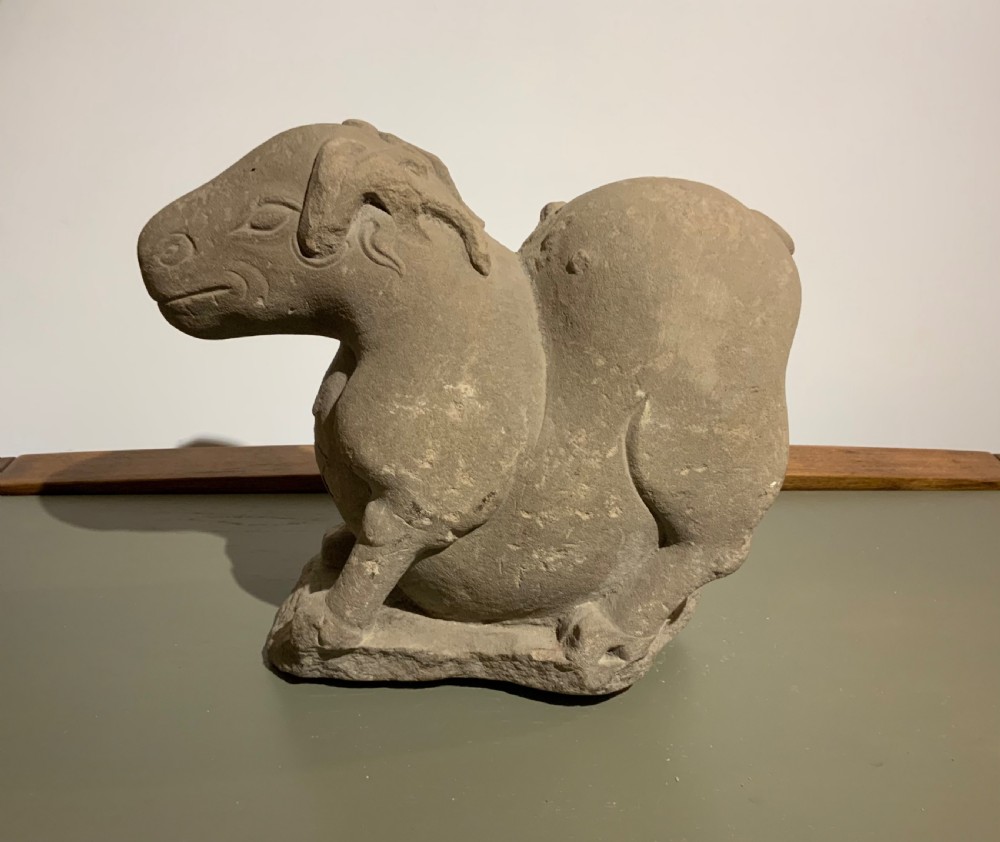 A carved warm grey stone sculpture depicting a resting ram with curled horns, crafted in an ancient Chinese style.