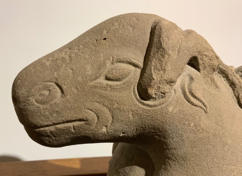 A carved warm grey stone sculpture depicting a resting ram with curled horns, crafted in an ancient Chinese style. View III