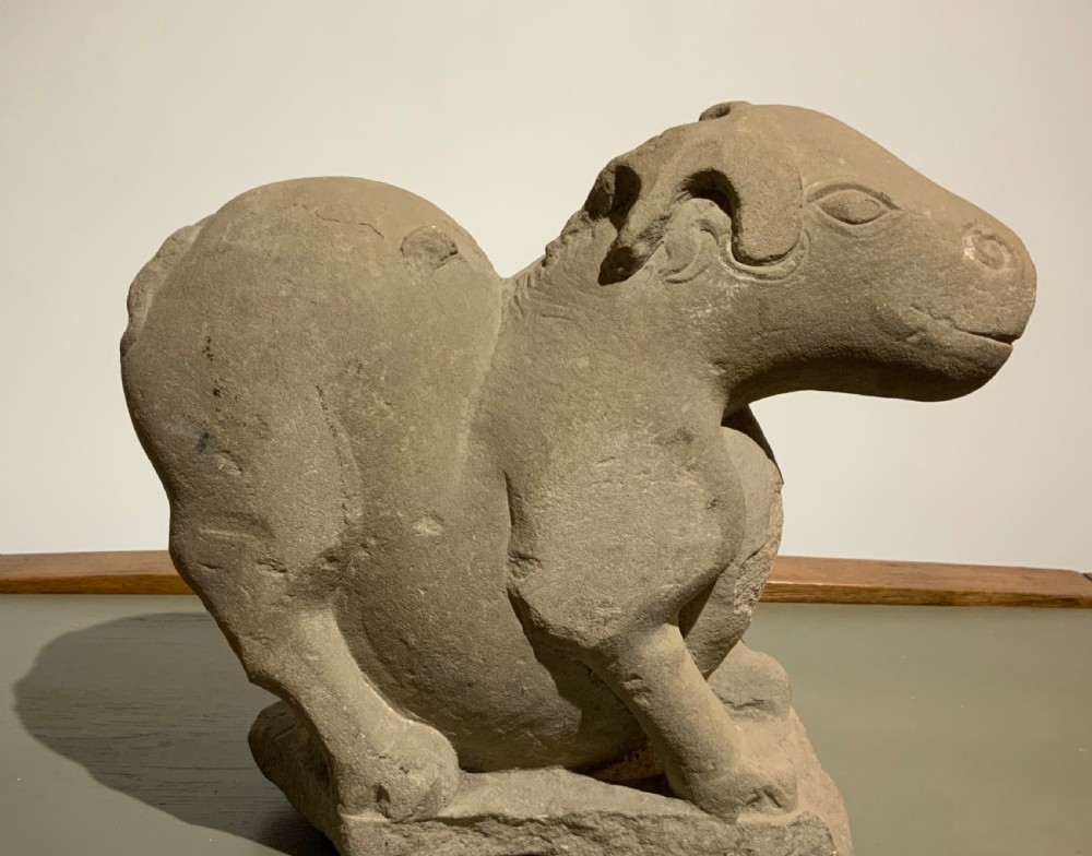 A carved warm grey stone sculpture depicting a resting ram with curled horns, crafted in an ancient Chinese style. View IV