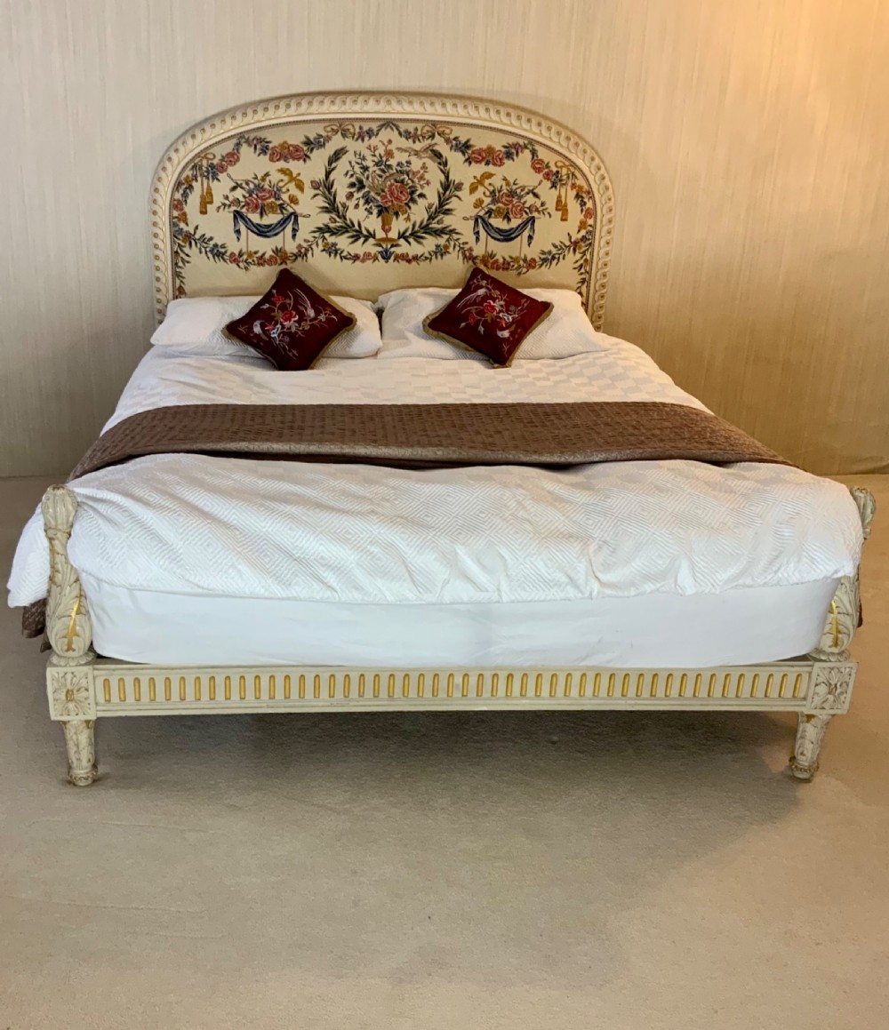 A carved cream painted and parcel gilt five-foot bed frame featuring a arched floral tapestry upholstered headboard and fluted footboard.